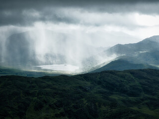 Lake surrounded by rolling hills with sunlight breaking through dark clouds and rain shafts.