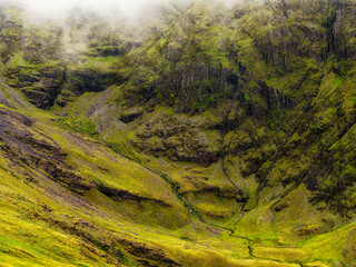 A verdant valley shows rocky cliffs and slopes blanketed in vibrant green moss and grass. A small stream courses through the valley floor.