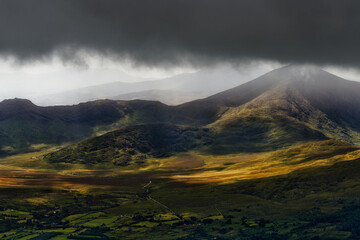 A view of a green, hilly landscape under dark storm clouds. Sunlight shines through the clouds, highlighting parts of the valley and the distant mountain peaks of County Kerry.