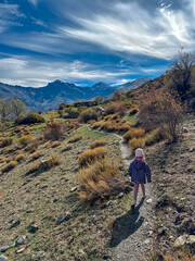 A young girl in a striped jacket walks on the rocky Vereda de la Estrella trail towards the majestic Mulhacen and Alcazaba peaks in the vast Sierra Nevada National Park, Andalusia, Spain.