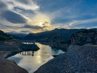 The calm waters of the Canales Reservoir reflect the golden sunset light near the Vereda de la Estrella, showing the intake tower and rugged Sierra Nevada mountains under a dramatic cloudy sky.