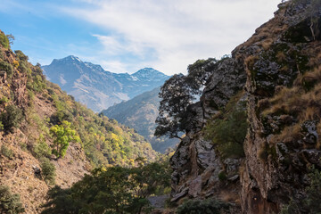 Steep rocky cliffs frame a dramatic view of the snow-capped Sierra Nevada peaks rising above the deep Genil valley, captured along the Vereda de la Estrella trail in Granada, Andalusia, Spain.