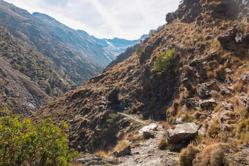 A scenic narrow rocky hiking trail winds along the steep slopes of the Genil valley, revealing the majestic snow-capped peaks of Sierra Nevada National Park in sunny Granada, Andalusia, Spain.