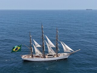 Aerial view of a three-masted sailing ship with sails and a large Brazilian flag on the stern sailing in the Atlantic Ocean. The training ship of the Brazilian Navy.