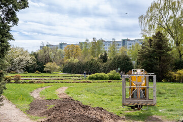 Urban park in spring with landscaped garden beds, green lawn and park maintenance equipment. Blooming trees (Salix sp., Betula sp.) and apartment blocks in background.