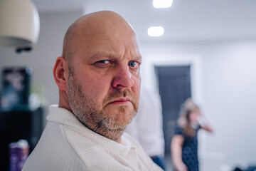 Bald man with a serious expression is looking directly at the camera in a modern indoor setting, with blurred figures in the background, conveying a sense of intensity and focus