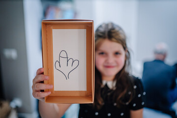 Young girl with brown hair, wearing a polka dot dress, is holding a box with a playful drawing inside, showcasing creativity and fun in a bright indoor setting