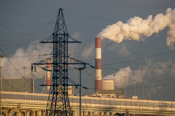 Industrial steam emissions come from a red-and-white chimney of a thermal power plant against the background of a power line pylon.