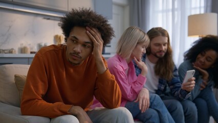 Stressed sad African American man sitting home frustrated male introvert headache ignoring friends while multiethnic people chatting mobile phone cellphone together social isolation gadget addiction