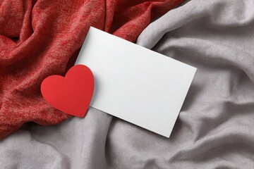A red heart rests on a white card, set against shimmering red and silver fabric, perfect for Valentines Day.