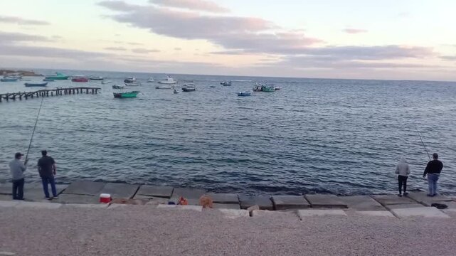 For fishing, fishermen are standing on rocks opposite Al-Kour Island (Miami Island). The island dates back to 30 BC. A great view of some boats, the Mediterranean Sea and the sky is near sunrise.