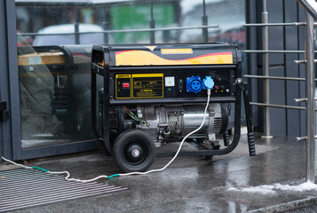 Portable black gasoline generator with connected power cables operating outside a store on a wet winter day.