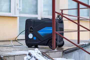 Portable power generator on outdoor stairs in winter, connected by cable near residential building, emergency electricity supply concept.