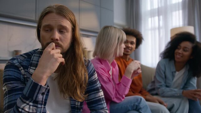 Upset worried Caucasian man thinking at home feeling lonely anxious excluded introvert male suffering stress ignoring friends while multiethnic women men having fun together social rejection isolation