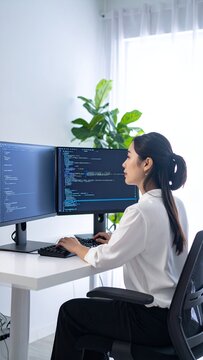 Focused Asian Woman Programmer Coding on Multiple Computer Screens in a Bright Office Environment.