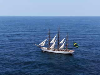 Fototapeta premium Aerial view of a three-masted sailing ship with sails and a large Brazilian flag on the stern sailing in the Atlantic Ocean. The training ship of the Brazilian Navy.
