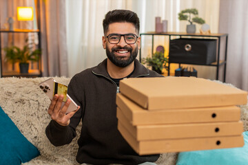 Indian man at home smiles holding smartphone and credit card while finishing online order and waiting for pizza delivery. Arabian guy on sofa enjoys food shopping, happy with purchase and fast service
