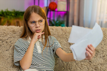 Child girl wears oversized mom's glasses, pretends to read important papers bills invoices on home sofa. School kid imitates serious adult conversation, acting confident expressive with funny gestures