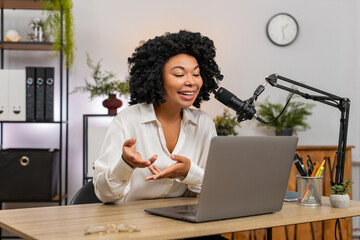 African American businesswoman at home office speaks into microphone gesturing recording podcast on laptop. Freelancer girl at table shares ideas smiling hoping loyal audience learns and channel grows