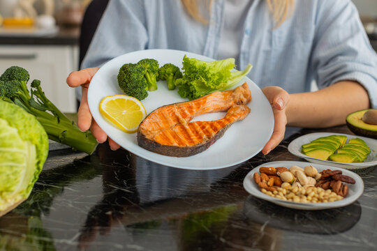 Woman holds plate with grilled salmon broccoli lettuce and lemon for keto lunch at home kitchen. Healthy low carb meal supports weight loss and clean eating with avocado and nuts on table for energy.