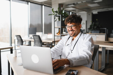 Fototapeta premium Smiling arab male doctor working on laptop in modern clinic office