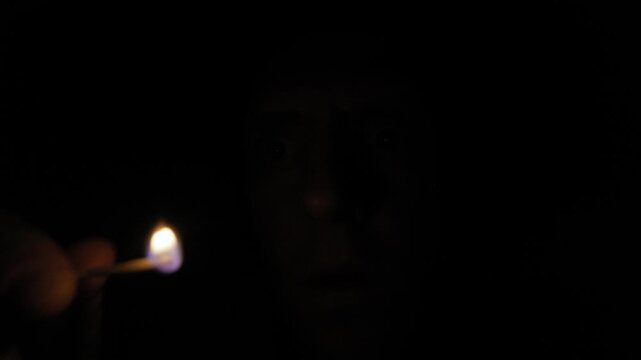 Man with a terrified expression lights a single match, revealing his face in the darkness. Extreme close-up uses dramatic chiaroscuro lighting against a pitch-black background for horror and suspense.