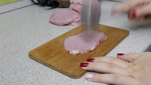 Female hands use a metal meat tenderizer to pound a raw pork chop on a wooden board. Close-up shot shows the process of preparing meat for cooking in a home kitchen.