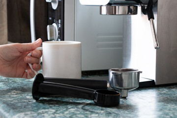 A hand holds a white ceramic mug near a coffee machine in a home kitchen. The process of making fresh coffee, morning ritual