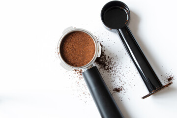 A metal coffee portafilter, measuring spoon, and loose coffee grounds lie on a white background