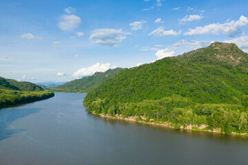 Fototapeta premium Expansive view of the Mekong river curving alongside densely forested green hills under a bright blue sky with scattered clouds. The scene features lush tropical vegetation, gentle slopes, and calm