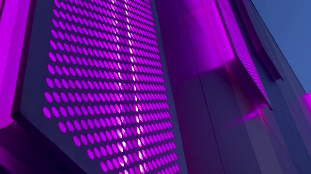 A close-up, low-angle view of a modern building facade featuring perforated purple panels that are dramatically illuminated at night, creating an abstract and futuristic architectural background.