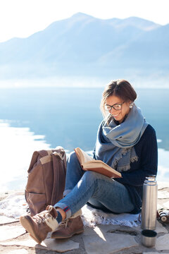 Happy woman reading book outdoors, relaxing in slow travel by mountain lake. Book adventure, self-development in nature, mindfulness, mental health in sabbatical. Digital detox, tech-free lifestyle