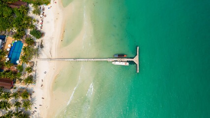 Aerial perspective shows a concrete pier extending from white sand into clear turquoise water at Coconut Beach, Koh Rong, Cambodia. Palm trees, a swimming pool, sun loungers, and anchored boats create © Florent