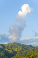 Fototapeta premium A towering white cloud rises above a sunlit green mountain ridge under a clear blue sky near Dien Bien Phu, Vietnam. The landscape is fresh and vibrant, with lush forest textures and soft evening
