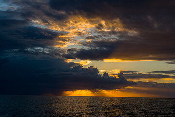 Obraz premium Heavy storm clouds gather above the Adriatic Sea as the sun sets, casting dramatic golden light and deep shadows across the water. The scene features bold contrasts and striking cloud formations over