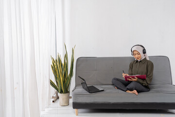 asian woman reading notebook on sofa, freelance writer journaling near laptop, wearing headphones, crosslegged and barefoot,