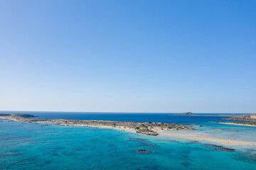 Sweeping aerial view of Elafonisi beach in Crete reveals a vivid blue lagoon, sandy sandbar, and rugged coastline beneath a vast, cloudless sky. The tranquil water and open horizon create a sense of