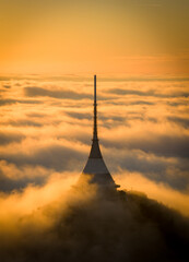 Aerial view of Je&scaron;těd Tower and Hotel rising above a sea of clouds at sunrise. Warm golden light and morning fog create a surreal, peaceful atmosphere over the Jizera Mountains in the Czech Republic.