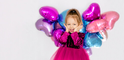 Fototapeta premium A banner dedicated to Valentine's Day, with a place to copy the text. A sweet happy little girl in a bright purple dress. Foil heart-shaped multicolored balloons