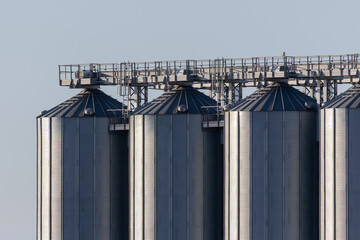 A quartet of industrial grain silos stand tall, linked by a steel framework under a clear sky. © Ardi
