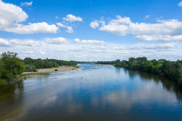 Fototapeta premium A broad stretch of the Loire river flows past a sandy bar and dense green forest beneath a sky scattered with fluffy cumulus clouds. The calm water reflects the blue sky and surrounding summer