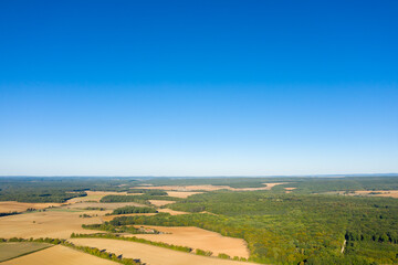 Obraz premium Expansive aerial view of golden farmland and dense green woodland stretching beneath a clear, vivid blue sky in the rural landscape of central France. The open fields and distant horizon create a