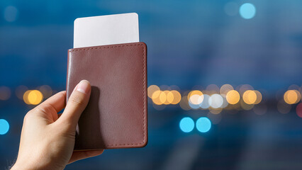 A man holds a leather-covered passport with a ticket inside. A blurred background (bokeh) of an evening airport or city with lights. The theme is travel, vacation, and departure.