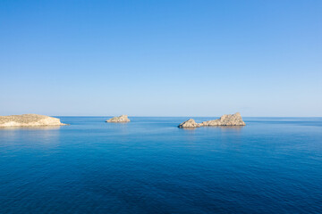 Obraz premium Three rugged rocky islets rise from the tranquil turquoise waters off the coast of Ambelou, Crete. The scene is bathed in clear morning light under a vast cloudless sky, evoking a sense of serenity