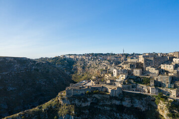 Fototapeta premium Wide view of Matera, Italy, featuring historic stone structures clustered atop a rugged plateau with steep cliffs, illuminated by clear daylight against a vivid blue sky.