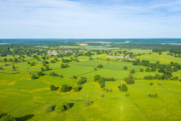 Obraz premium Aerial view of expansive green fields divided by hedgerows and dotted with trees, with a small village nestled in the distance under a bright blue sky near Cuncy les Varzy.