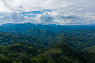 Obraz premium Vast mountain ridges covered in dense green vegetation stretch into the distance beneath a sky filled with towering cumulus clouds in the rural region between Phonsavan and Phou Khoun, Laos. The