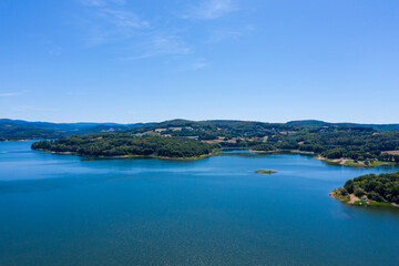 Obraz premium Wide view of Panneciere-Chaumard reservoir with deep blue water, surrounded by rolling forested hills and scattered farmland under a clear summer sky in Burgundy. The composition highlights tranquil