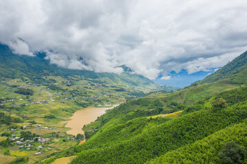 Fototapeta premium Sweeping view of a muddy river winding through lush green terraced rice fields and scattered rural homes in the Sapa valley, Vietnam. Dramatic low clouds hang over the mountainous landscape, creating