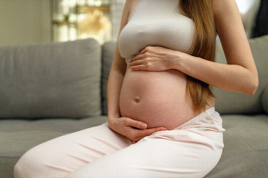 Pregnant woman sits on sofa, cradling her belly with both hands, showing anticipation and maternal love - Powered by Adobe